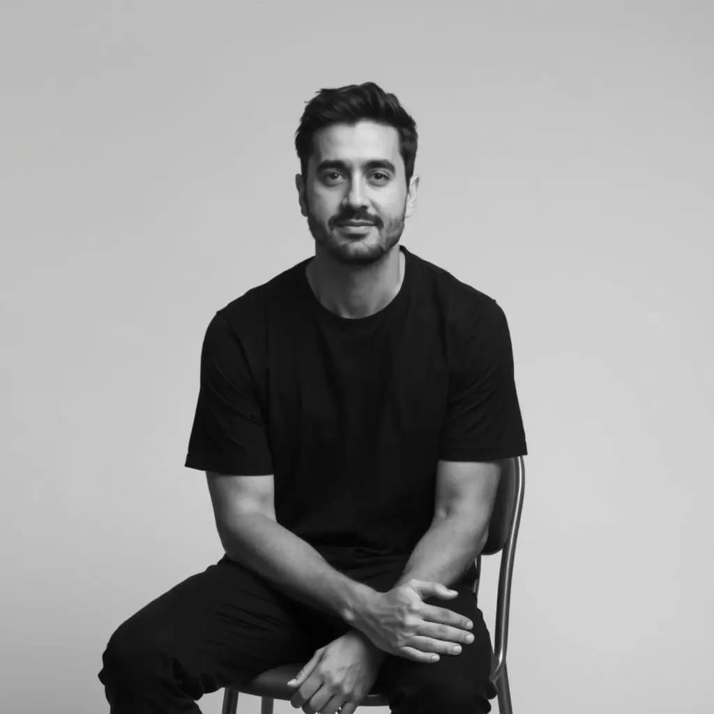 صمّم صورة شخصية بأسلوب فوتوغرافي بسيط ومركّز، بالأبيض والأسود لملحم زين يجلس على كرسي بزاوية أمامية، يرتدي تيشيرت أسود وبنطال داكن. الإضاءة ناعمة وموزعة بشكل متوازن لتُبرز ملامح الوجه وتفاصيل الملابس دون ظلال قاسية. الخلفية بيضاء ناعمة وخالية من التفاصيل، ما يمنح التركيز الكامل على تعبير الوجه